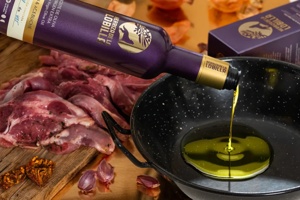 Aceite de acebuche: El olivo silvestre y ancestral. Conoce 4 de sus mayores beneficios. 8 Botella de aceite de oliva virgen extra variedad lechín y acebuche vertiendo el producto sobre una sartén con una tabla de carne macerada al fondo