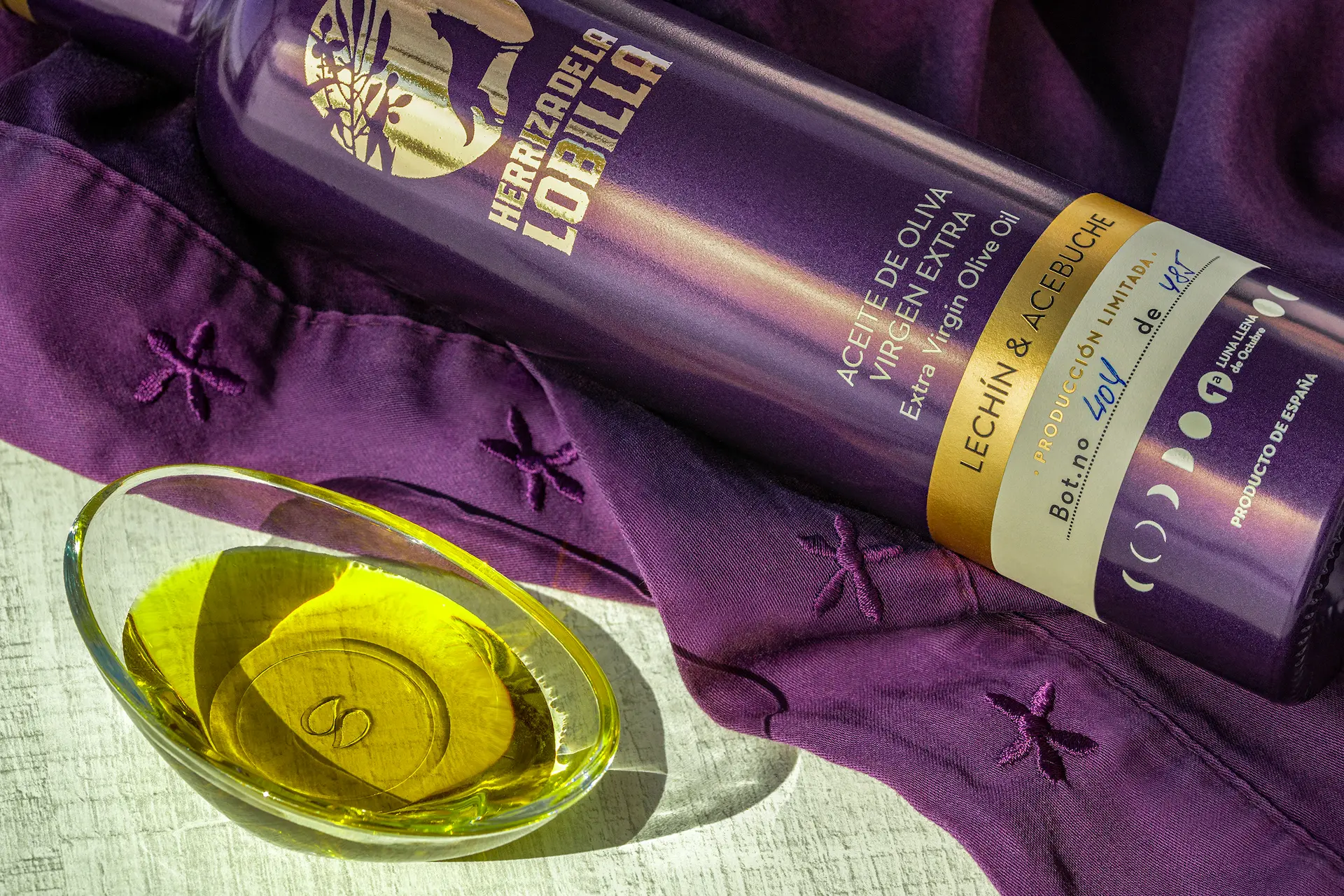 Botella de aceite de oliva virgen extra variedad lechín y acebuche tumbada, mostrando el código único de edición limitada y una muestra del producto sobre un recipiente de cristal