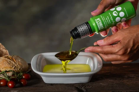 Botella pequeña de aceite de oliva virgen extra variedad hojiblanca vertiendo el producto sobre una cuchara y un recipiente con fondo tipo bodegón y pan rústico